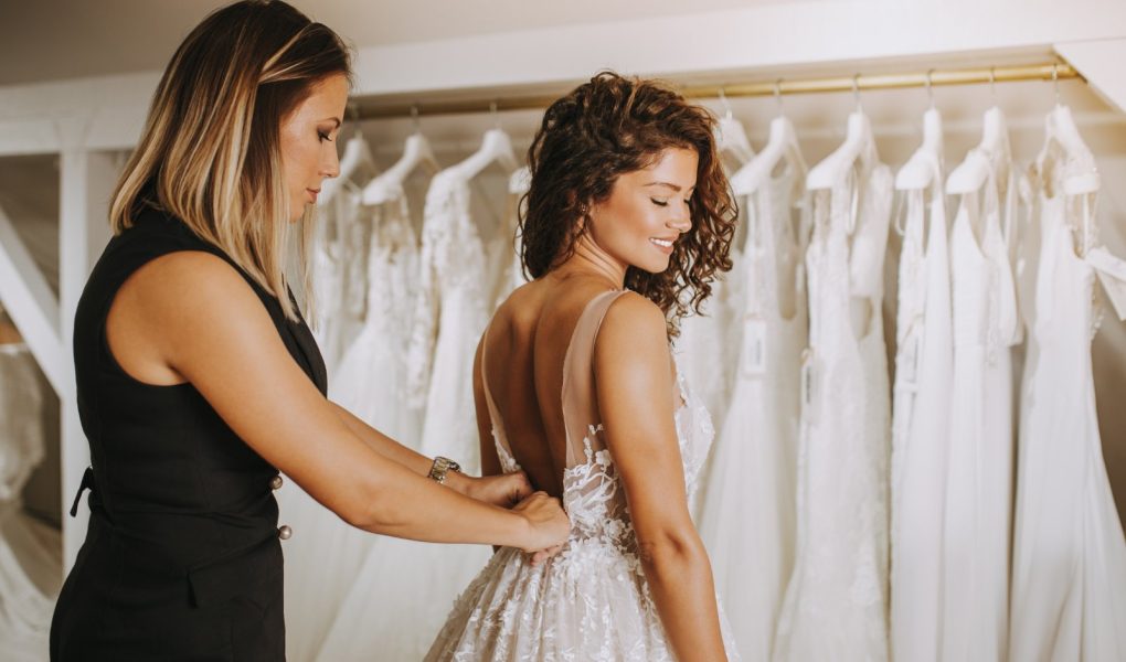 Future mariée essayant sa robe de mariée en boutique, aidée par une conseillère
