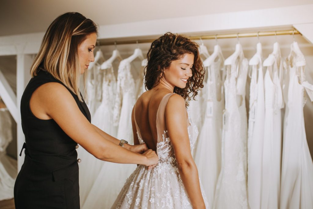 Future mariée essayant sa robe de mariée en boutique, aidée par une conseillère