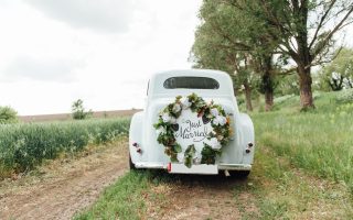 Voiture de mariage ancienne blanche décorée d'une couronne de fleurs sur un chemin de campagne