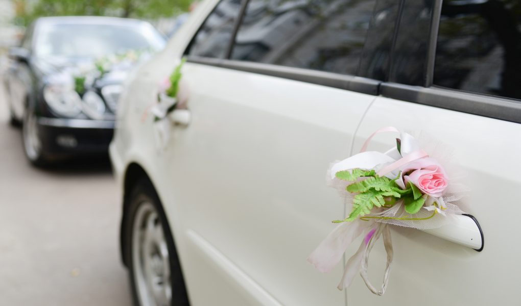Cortège de mariage : voiture blanche décorée de fleurs et de rubans, suivie d'une voiture noire
