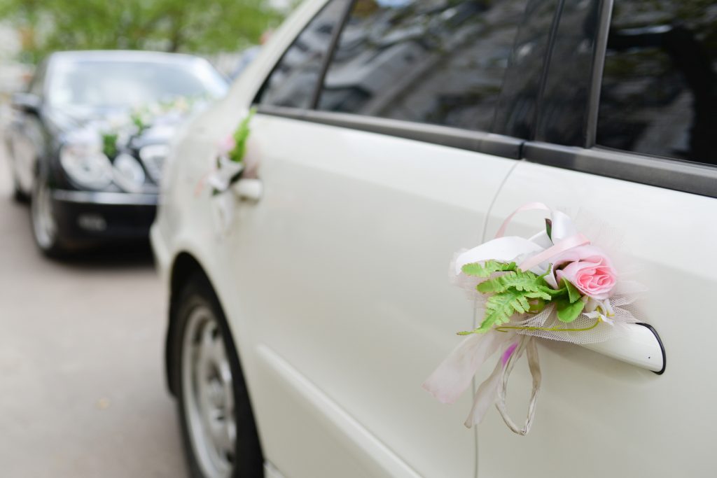 Cortège de mariage : voiture blanche décorée de fleurs et de rubans, suivie d'une voiture noire