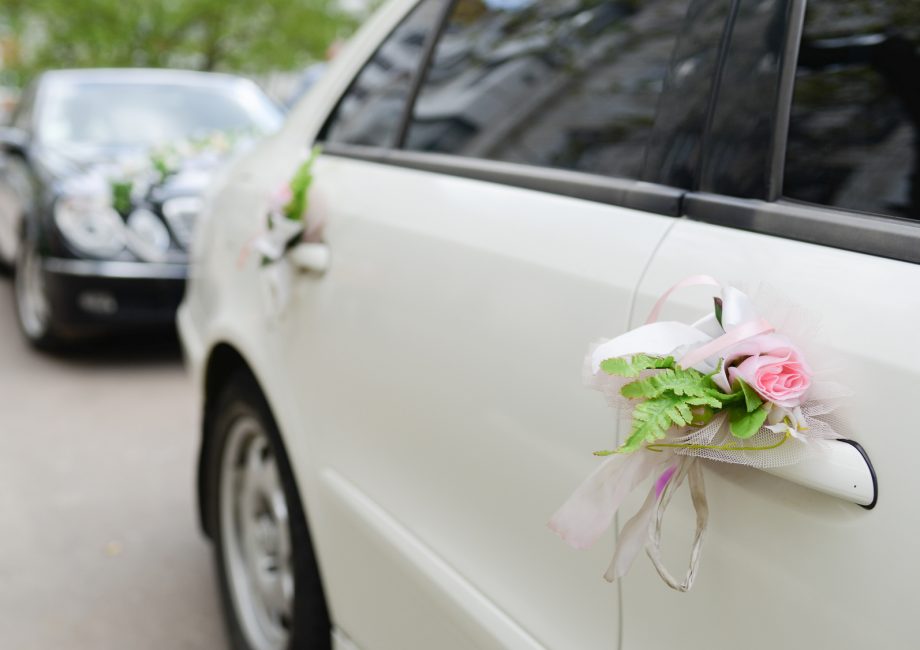 Cortège de mariage : voiture blanche décorée de fleurs et de rubans, suivie d'une voiture noire