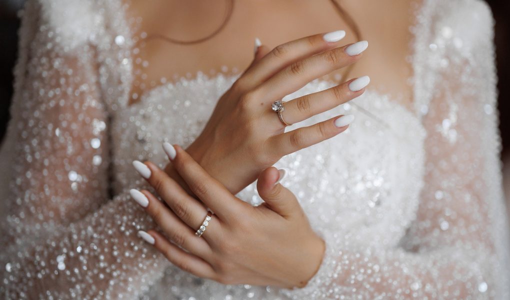 Mains de mariée aux ongles blancs manucurés, avec bague de fiançailles et alliance, sur une robe scintillante