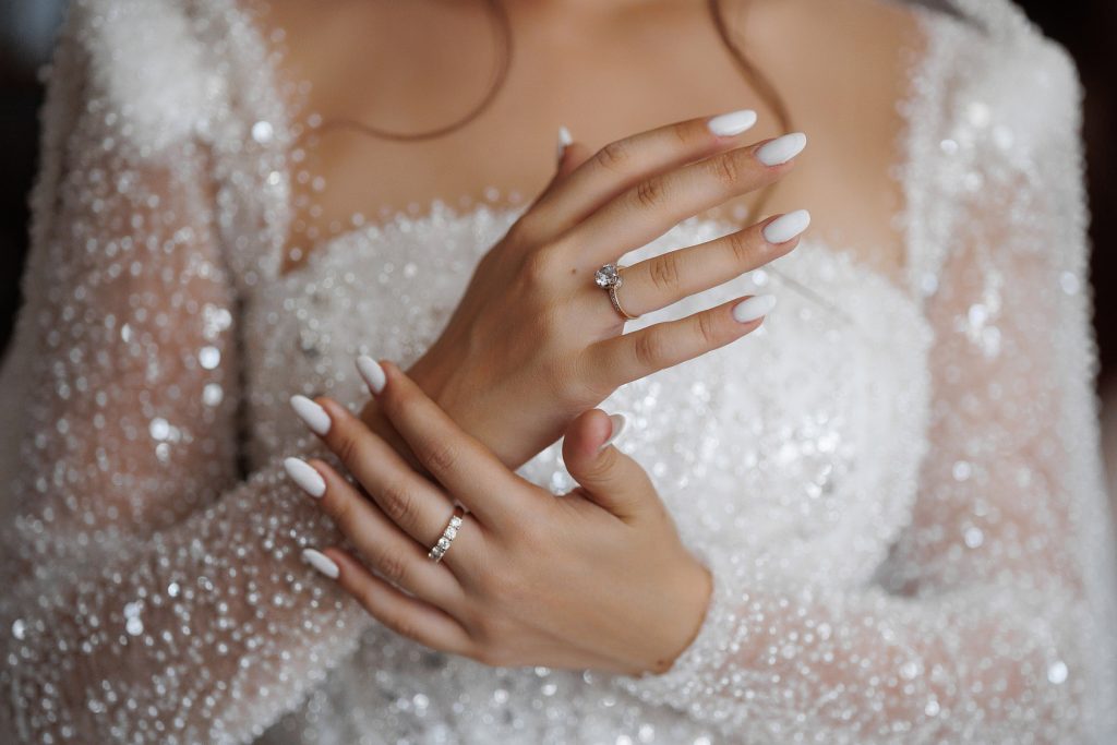 Mains de mariée aux ongles blancs manucurés, avec bague de fiançailles et alliance, sur une robe scintillante
