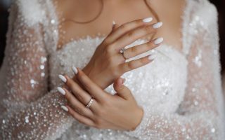 Mains de mariée aux ongles blancs manucurés, avec bague de fiançailles et alliance, sur une robe scintillante
