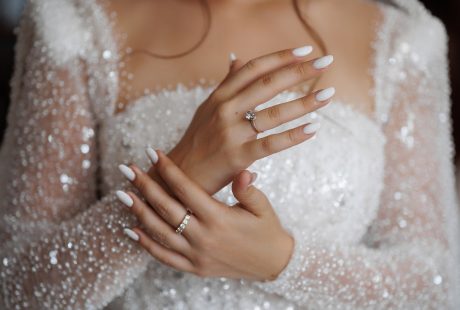 Mains de mariée aux ongles blancs manucurés, avec bague de fiançailles et alliance, sur une robe scintillante