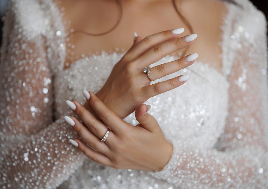 Mains de mariée aux ongles blancs manucurés, avec bague de fiançailles et alliance, sur une robe scintillante