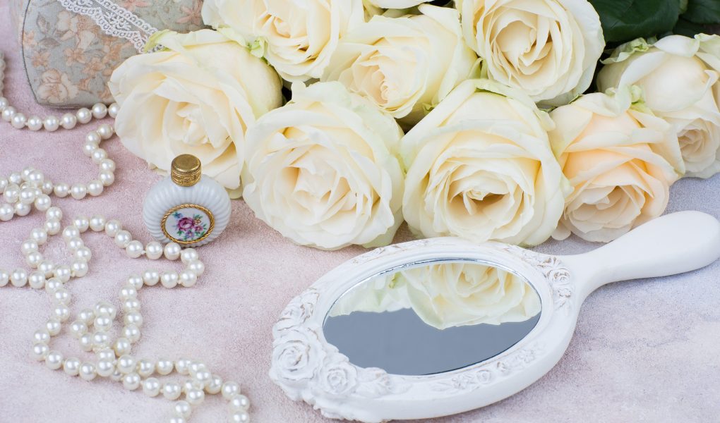 Flacon de parfum, perles et bouquet de roses blanches devant un miroir