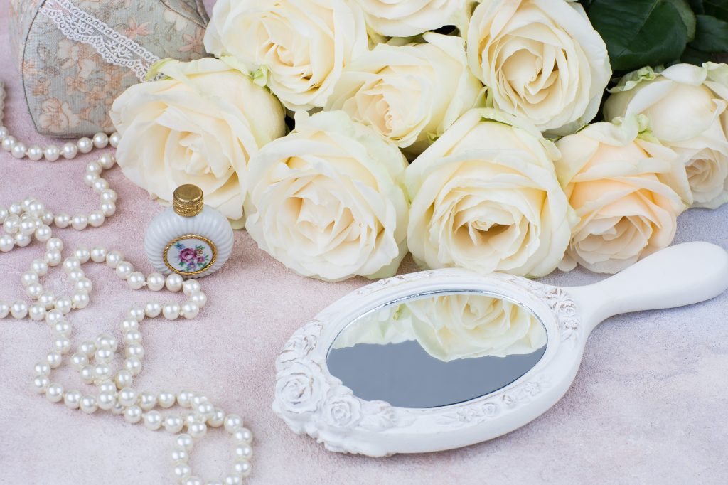 Flacon de parfum, perles et bouquet de roses blanches devant un miroir